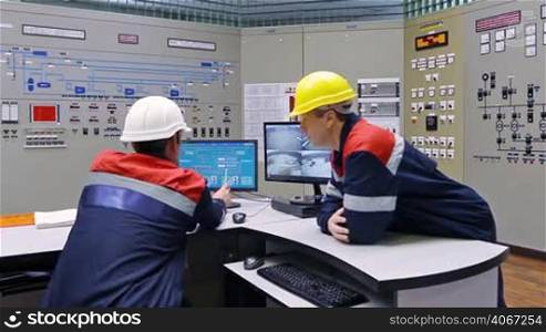 two engineers talk while looking at circuit and diagrams in monitors near main control panel of gas compressor station