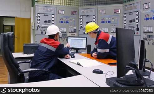 two engineers talk while looking at circuit in monitor near main control panel of gas compressor station