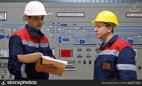 two engineers talk while looking into logs and giving explanation about circuit at main control panel of gas compressor station, closeup