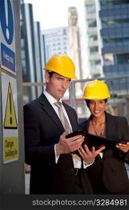 Two executives, one man and one women, wearing hard hats review plans in a modern city environment. The focus is primarily on the man in the foreground.