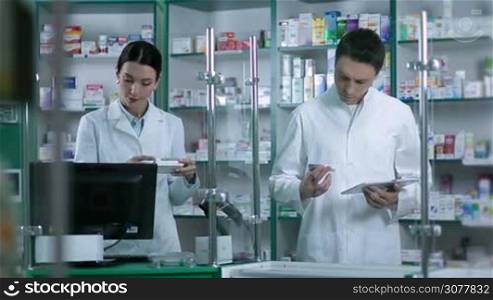 Two experienced pharmacists working in pharmacy. Confident male druggist checking medicines while using touchpad. Beautiful brunette chemist woman holding drugs and typing on computer keyboard. Health care and pharmacology concept.