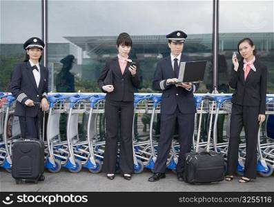 Two female cabin crews holding mobile phones and standing with two pilots at an airport