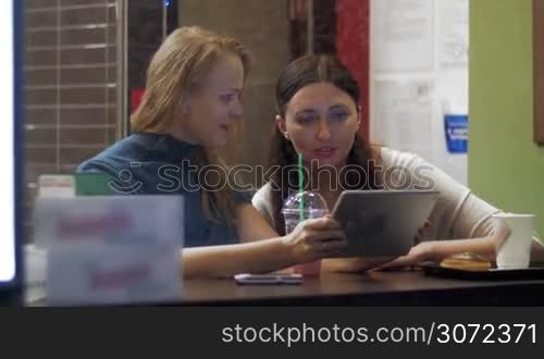 Two female friends are sitting in cafe in rainy day and watching photos in tablet PC. Shot is made through the cafe show window.