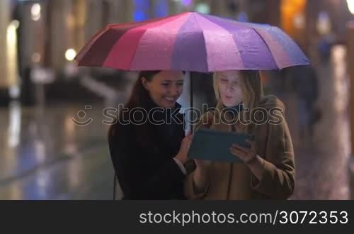 Two female friends are standing under an umbrella and watching something funny in tablet PC.
