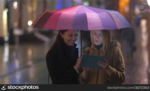 Two female friends are standing under an umbrella and watching something funny in tablet PC.