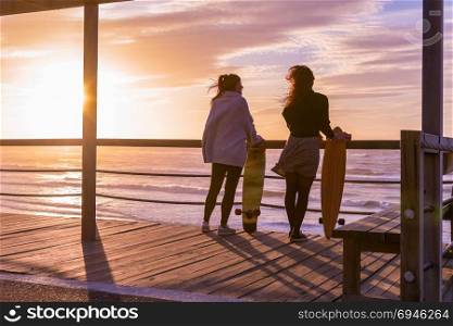 Two female friends chatting with skateboard near the beach at sunset.