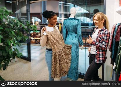 Two female friends choosing dresses in shop, shopping. Shopaholics in clothing store, consumerism lifestyle, fashion. Two female friends choosing dresses, shopping