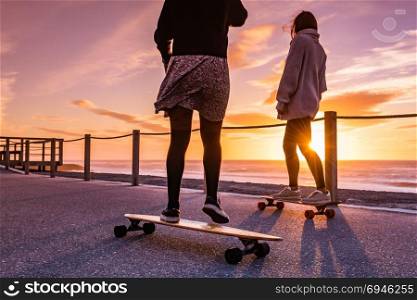 Two female friends playing with skateboard near the beach at sunset.