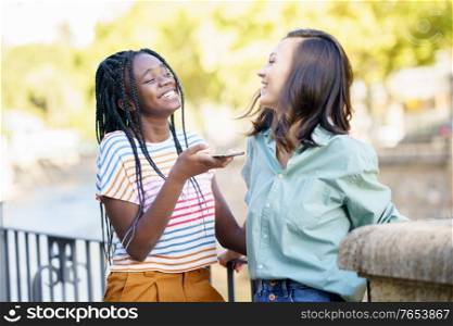 Two female friends recording a voice note with smartphone outdoors. Multiethnic friends.. Two female friends having fun together on the street. Multiethnic friends.