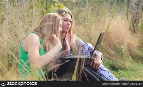 Two female friends sitting on the grass and talking online using laptop.