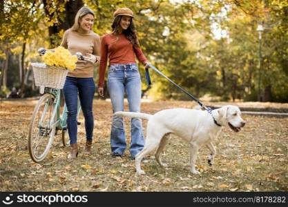 Two female friends walking in the yellow autumn park with dog and bicycle