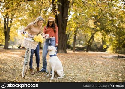 Two female friends walking in the yellow autumn park with dog and bicycle