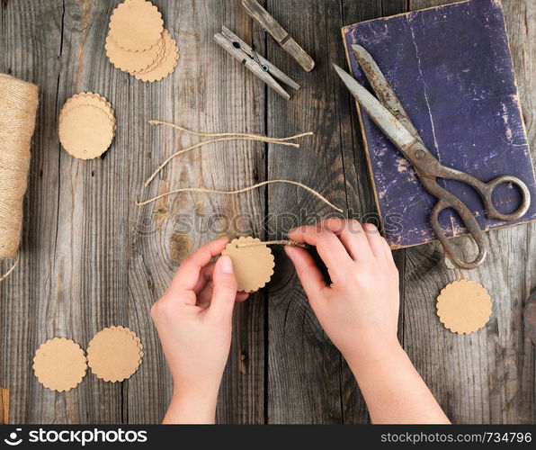 two female hands and paper tags with a brown rope on a gray wooden table, lesson on the manufacture of price tags, top view