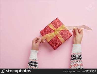 two female hands are holding a red box wrapped in gold ribbon on a pink background, top view. Holiday greetings concept
