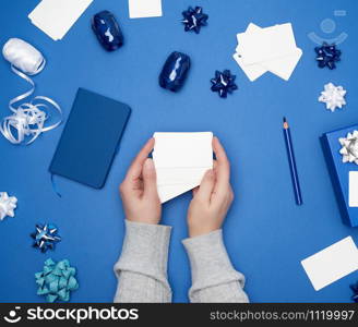 two female hands are holding white paper blank business cards, on a blue background are wrapped gifts and decor, top view