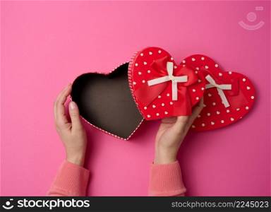 two female hands hold a red heart-shaped box on a pink background. Festive backdrop
