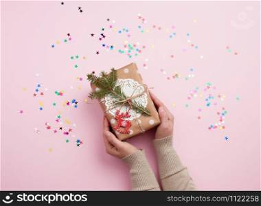 two female hands holding a gift wrapped in paper and decorated with a rope and a sprig of spruce on a pink background, top view