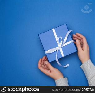 two female hands in a sweater hold a square blue box with a bow on a blue classic background, top view, festive backdrop