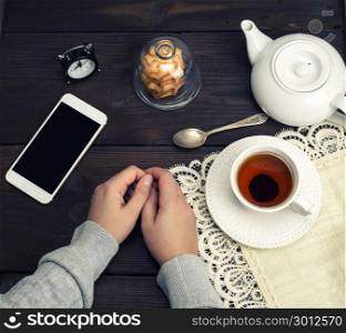 two female hands lie on a wooden table, next to a cup of tea and a smartphone with a black screen, top view