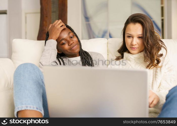 Two female student friends sitting on the couch at home using a laptop. Multiethnic women.. Two female student friends sitting on the couch at home using a laptop.