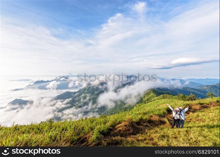 Two female tourists, mother and daughter stand waving amidst the beautiful natural landscape of mountain range sky cloud and fog in the winter at Phu Chi Fa Forest Park, Chiang Rai, Thailand