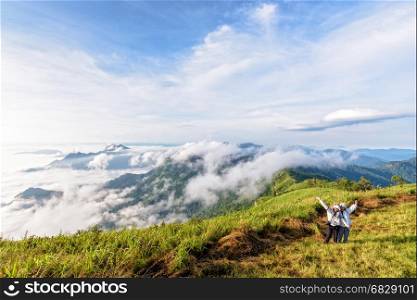 Two female tourists, mother and daughter stand waving amidst the beautiful natural landscape of mountain range sky cloud and fog in the winter at Phu Chi Fa Forest Park, Chiang Rai, Thailand
