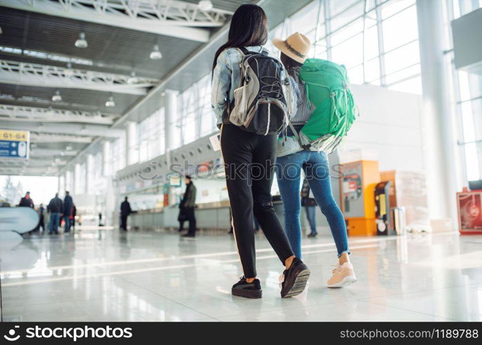 Two female tourists with backpacks in waiting area of international airport. Passengers with baggage in air terminal, back view, happy journey of white and black ladies, summer travel of happy women. Female tourists in waiting area of airport