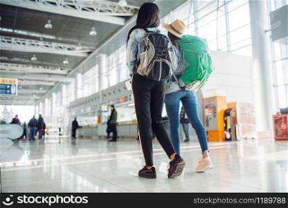 Two female tourists with backpacks in waiting area of international airport. Passengers with baggage in air terminal, back view, happy journey of white and black ladies, summer travel of happy women. Female tourists in waiting area of airport