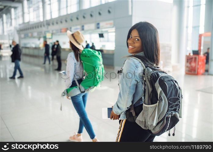 Two female tourists with backpacks in waiting area of international airport. Passengers with baggage in air terminal, back view, happy journey of white and black ladies, summer travel of happy women