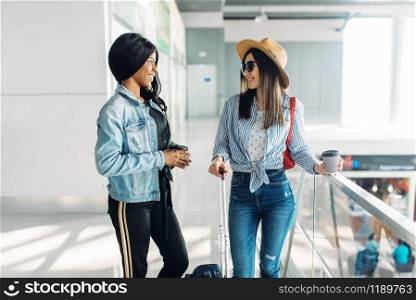 Two female travelers with luggage begins to travel in airport. Passengers with baggage in air terminal, back view, happy journey of white and black ladies, summer travel. Two female travelers with luggage in airport