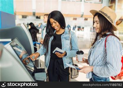 Two female travelers withdraw cash at ATM in airport. Passengers with baggage in air terminal, back view, happy journey of white and black ladies, summer travel