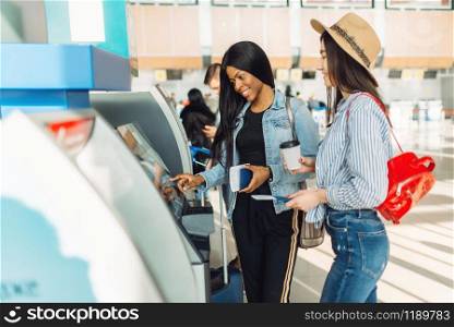 Two female travelers withdraw cash at ATM in airport. Passengers with baggage in air terminal, back view, happy journey of white and black ladies, summer travel. Female travelers withdraw cash at ATM in airport