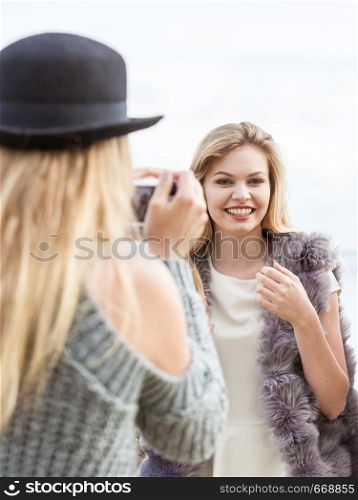 Two females friends having fun during outdoor photo session. Woman taking pictures of two during warm autumn weather.. Photo shoot of fashion models