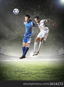 two football players in jump to strike the ball at the stadium under rain