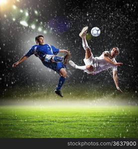 two football players in jump to strike the ball at the stadium under rain