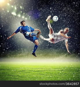 two football players in jump to strike the ball at the stadium under rain