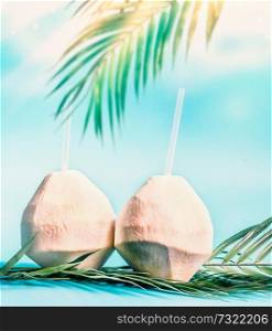 Two fresh coconut cocktails with tropical leaves at blue sky background with hanging palm leaves and sunshine. Tropical vacation. Summer holiday. Beach party. Healthy natural coconut water drink