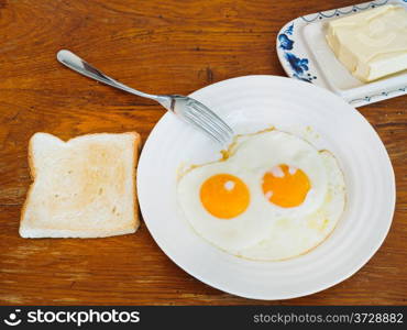 two fried eggs on white plate, fresh toast and dairy butter in butterdish on wooden table