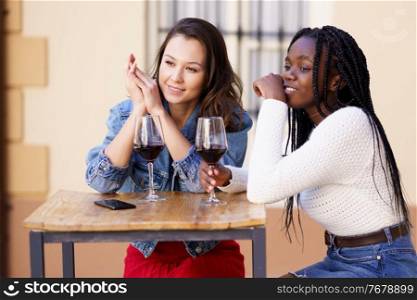 Two friends drinking red wine sitting at a table outside a bar. Multiethnic women.. Two women drinking red wine sitting at a table outside a bar.