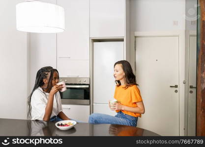 Two friends having a healthy snack while chatting at home. Multiethnic women.. Two friends having a healthy snack while chatting at home