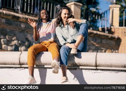 Two friends having fun together on the street sitting on a urban wall. Multiethnic women.. Two friends having fun together on the street sitting on a urban wall.