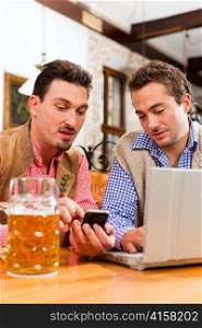 Two friends in Bavarian pub in traditional clothes sitting on a table in a pub with laptop