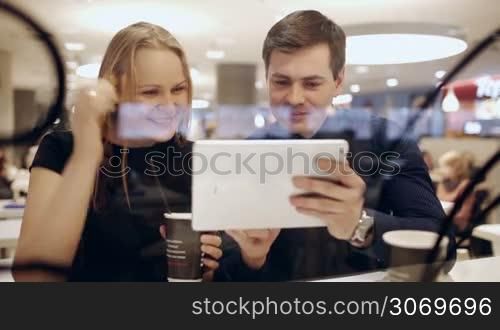 Two friends or colleagues in cafe having exctiting talk about something shown on pad. Woman drinking coffee. View through the glass