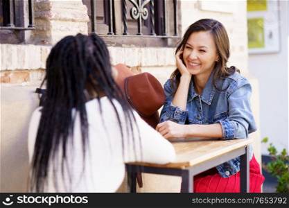 Two friends talking sitting at a table outside a bar. Multiethnic women.. Two multiethnic friends talking sitting at a table outside a bar.