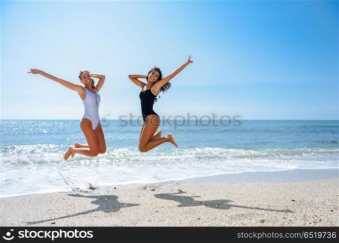 Two funny girls in swimsuit jumping on a tropical beach. Two funny girls with beautiful bodies in swimwear jumping on a tropical beach. Funny caucasian and arabic females wearing black and white swimsuits.
