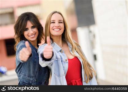 Two funny women friends with thumbs up and looking at the camera in urban background. Young girl wearing casual clothes.