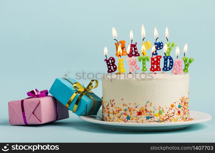 Two gift boxes near the cake with happy birthday candles against blue backdrop
