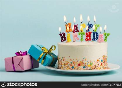 Two gift boxes near the cake with happy birthday candles against blue backdrop