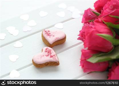 two gingerbread hearts on a wooden background, confetti hearts and a bouquet of bright pink roses.
