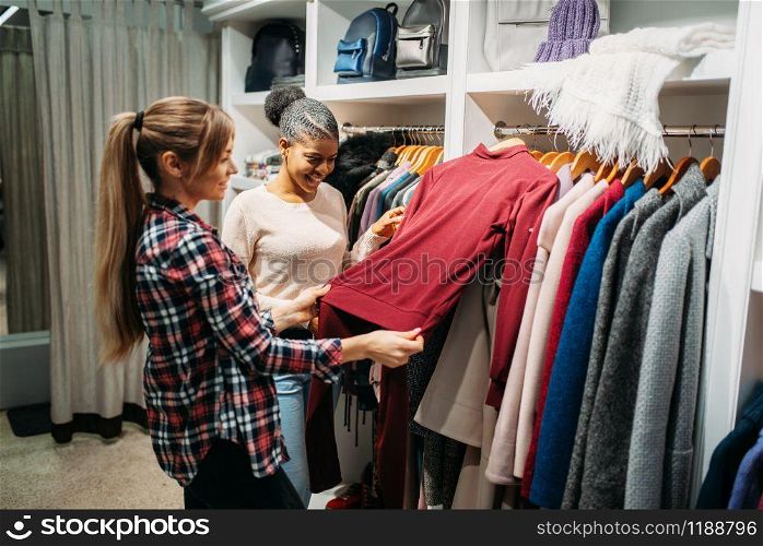 Two girlfriends choosing clothes in shop, shopping. Shopaholics in clothing store, consumerism lifestyle, fashion